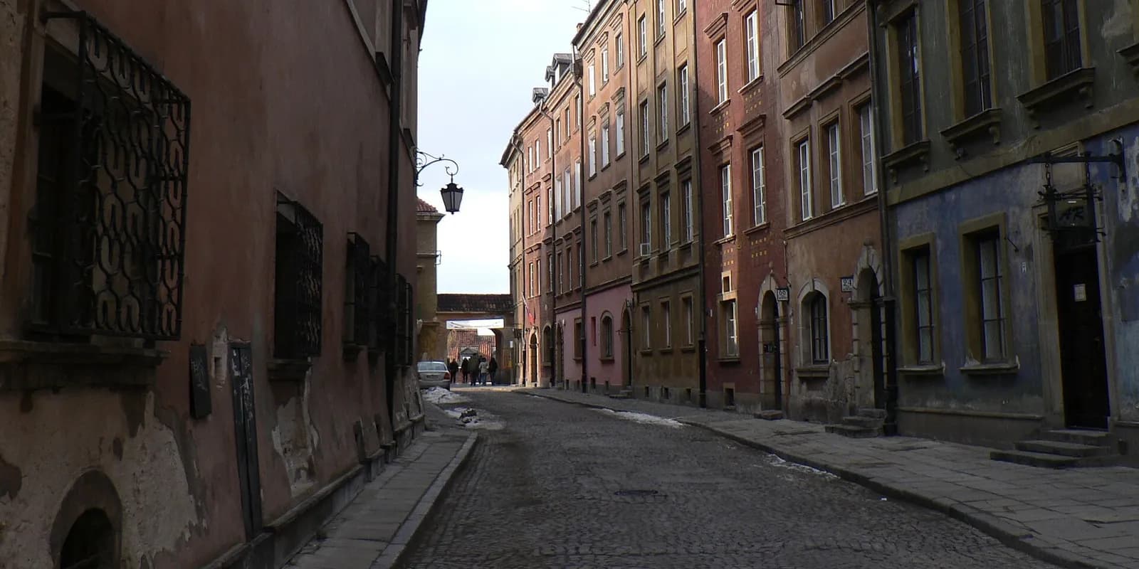 A narrow cobblestone street is lined with tall, old buildings in various shades of red, orange, and blue under an overcast sky.