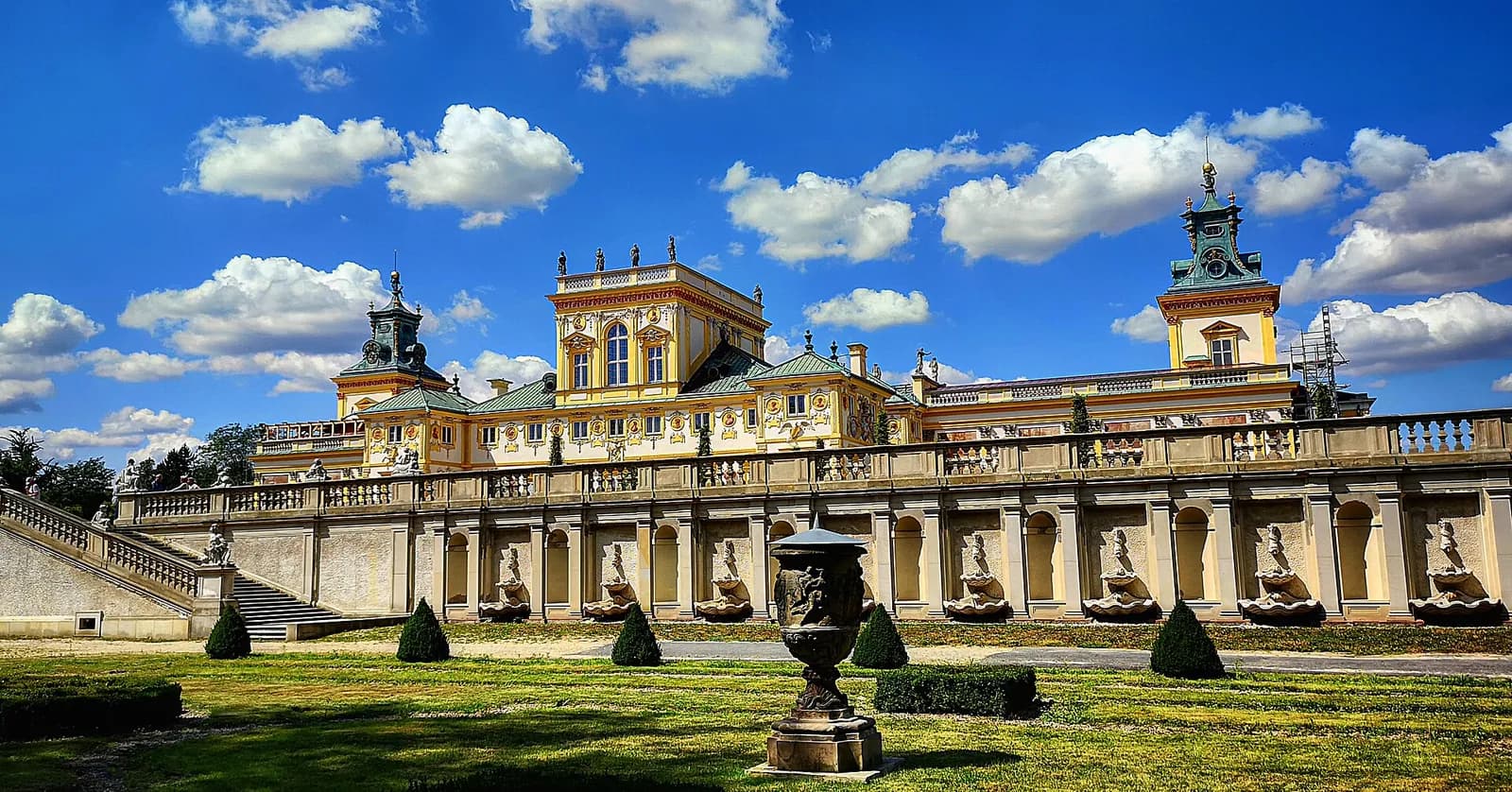 The Wilanów Palace in Warsaw, Poland is a grand yellow and white building with a green roof, set against a bright blue sky with fluffy white clouds, and surrounded by a manicured garden with a large stone staircase and small evergreen shrubs.