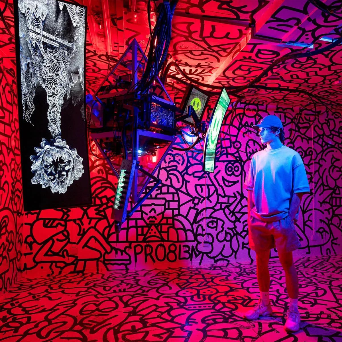 A man in a blue cap stands in a room covered in black doodle-like graffiti on a red and pink background, with glowing electronic equipment and a white abstract art piece on the left.