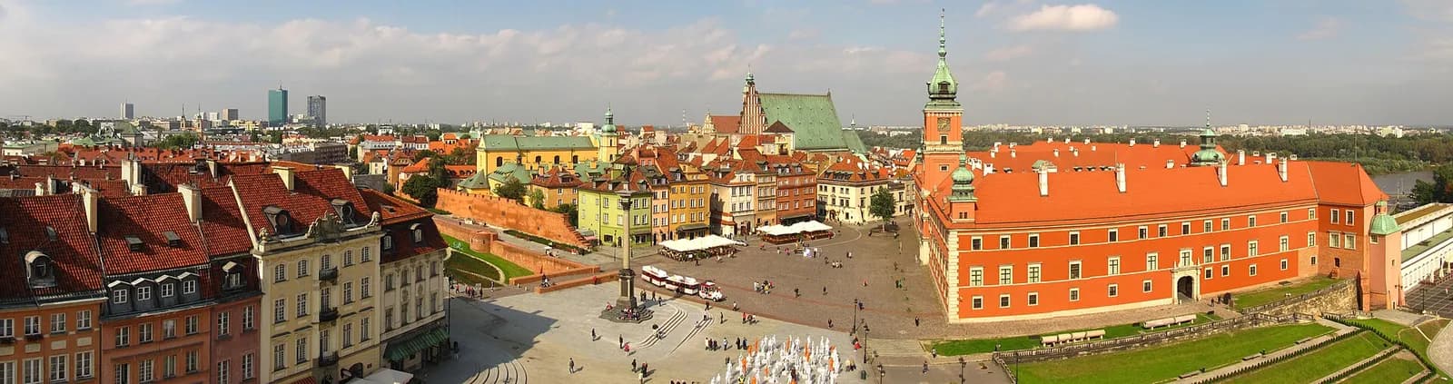 A panoramic aerial view of Warsaw's Old Town showing a large square with people, surrounded by buildings with orange roofs, including the Royal Castle and St. John's Archcathedral, under a clear sky.