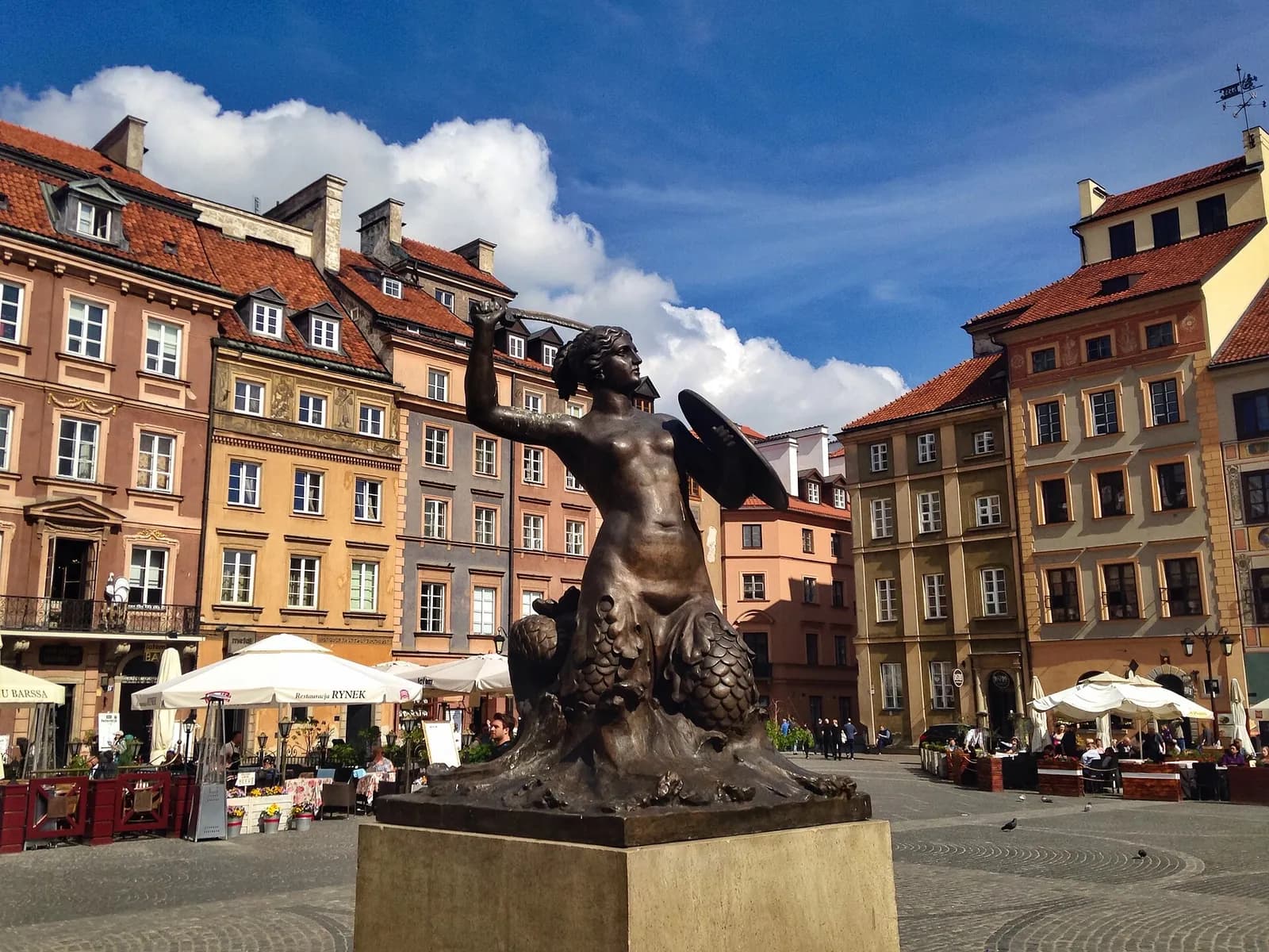 A bronze mermaid statue stands prominently in a bustling town square, surrounded by colorful buildings with red-tiled roofs under a blue sky with white clouds.