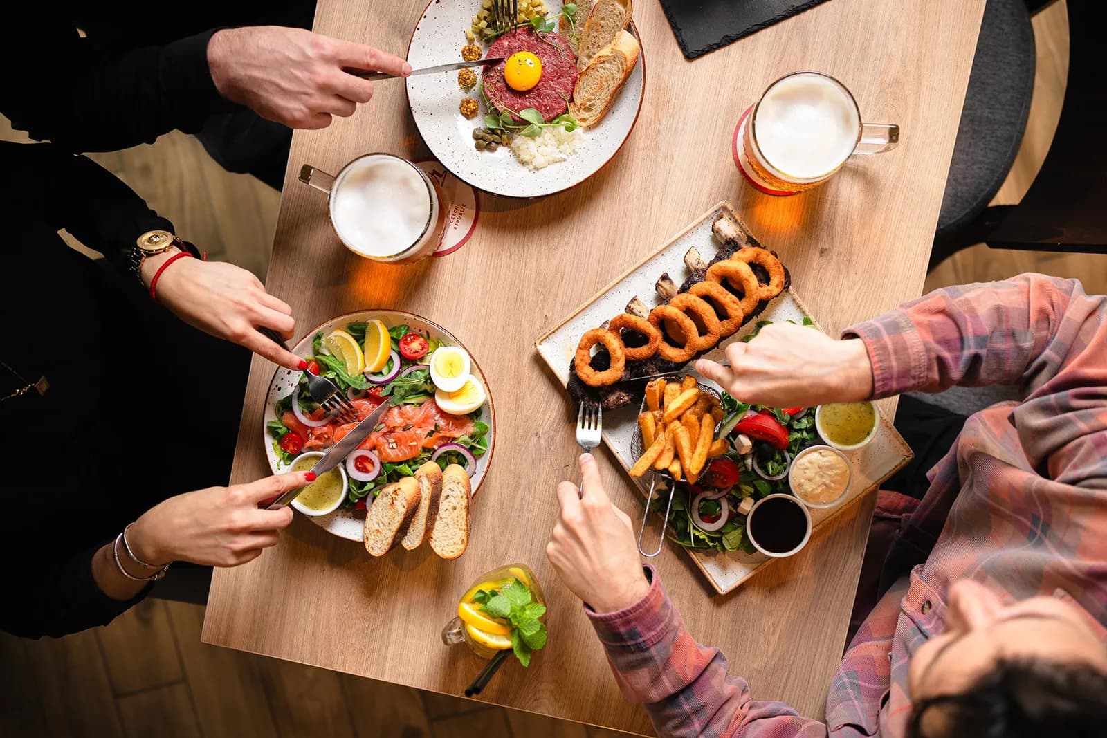 A top-down view of people sharing a meal with steak tartare, salmon salad, and ribs with onion rings and fries, alongside glasses of beer and a lemon drink.