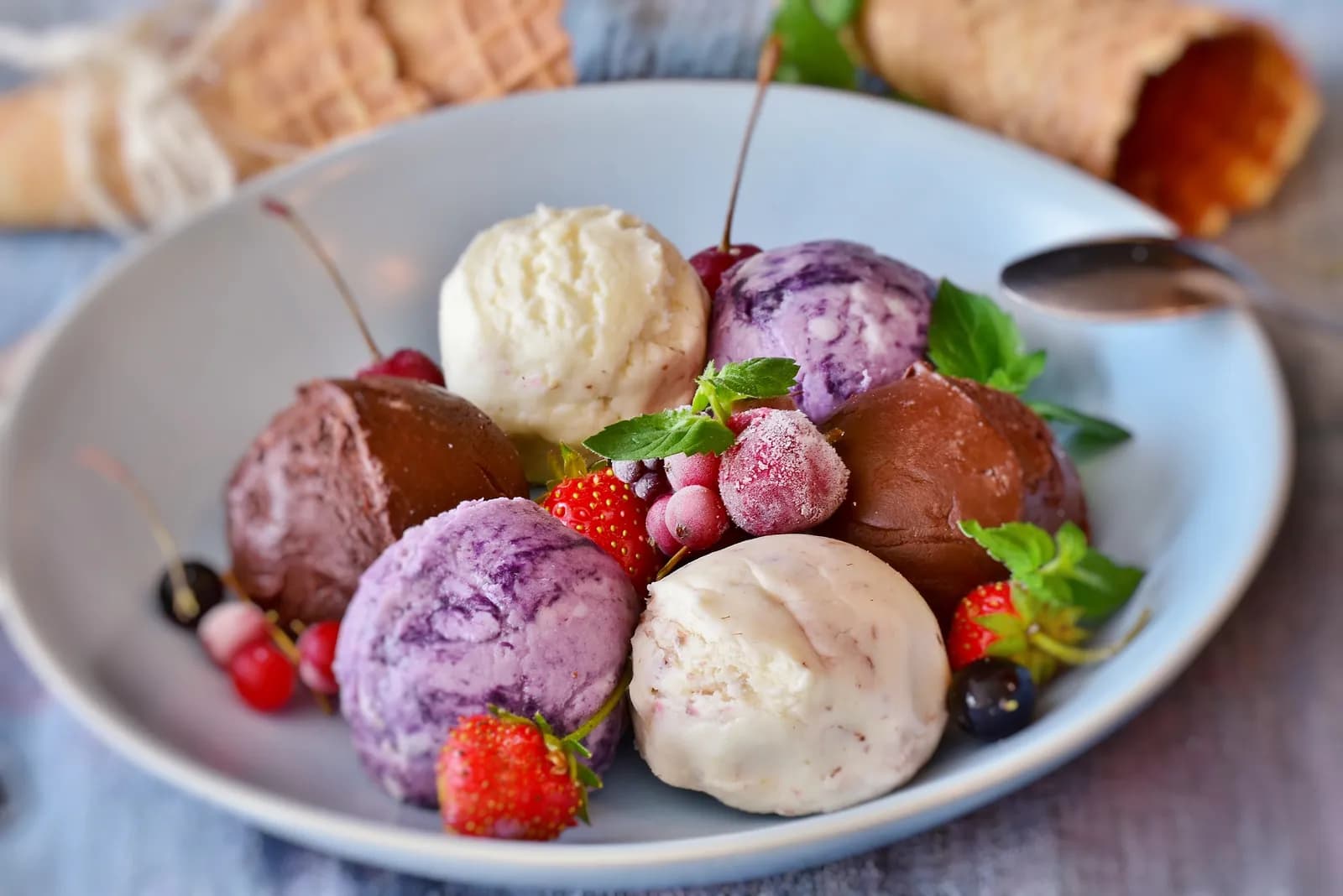 A light blue bowl filled with multiple scoops of ice cream in various colors and flavors, garnished with fresh berries and mint leaves.