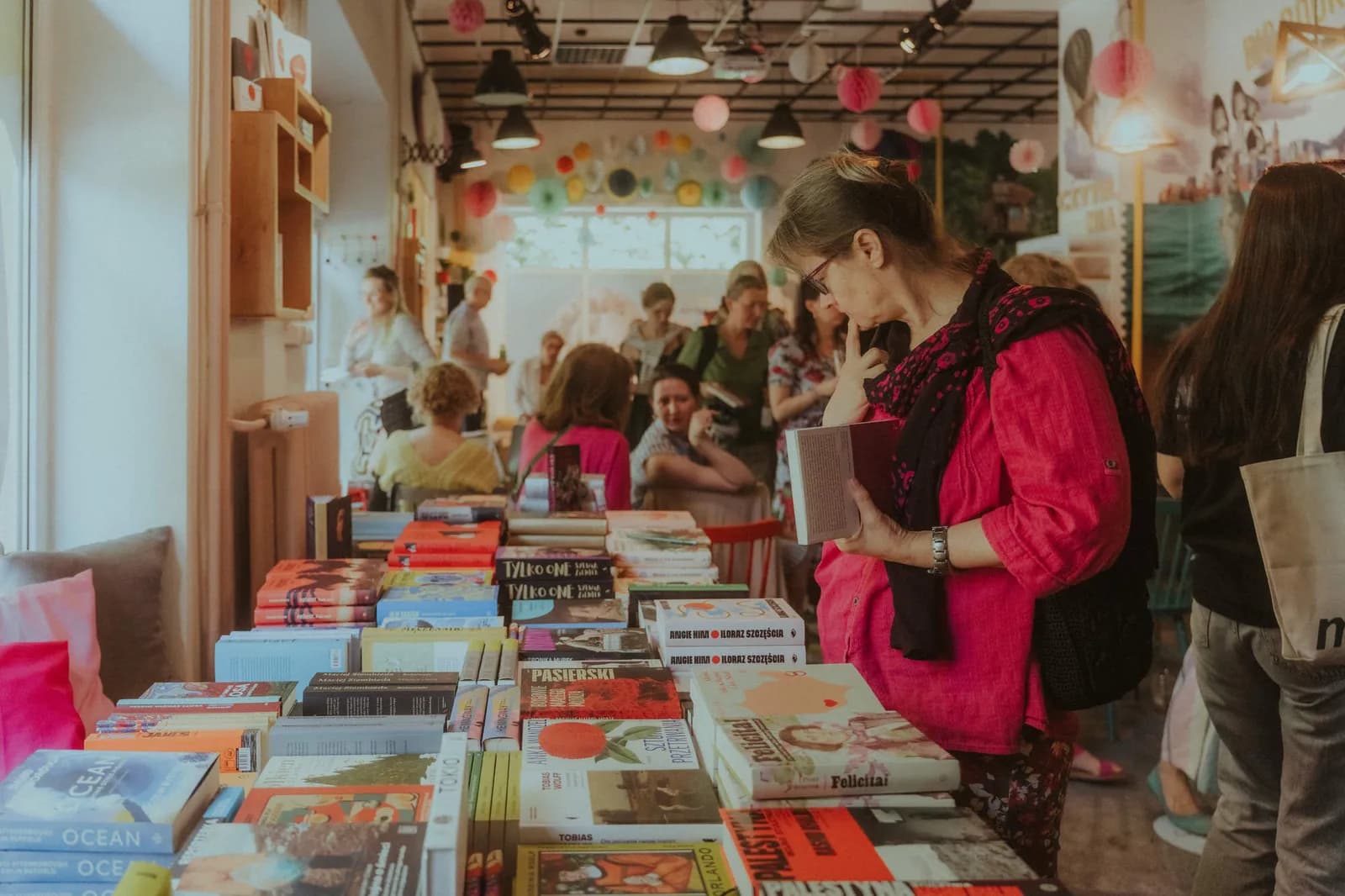 A woman in glasses and a pink shirt holds an open book while browsing a large selection of books displayed on tables in a busy indoor market.
