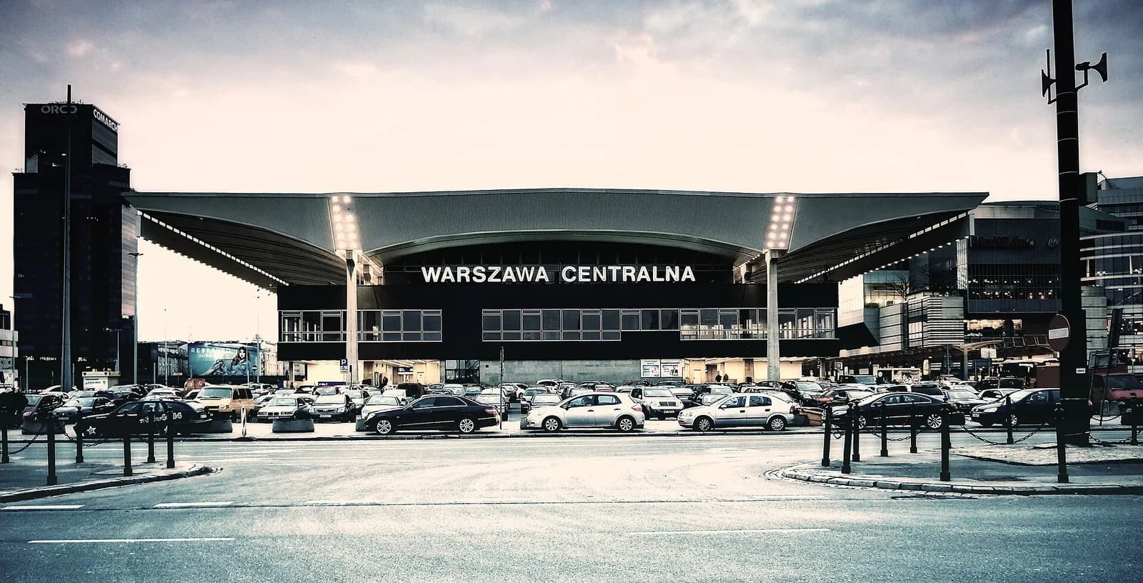 The Warszawa Centralna train station building with its distinctive curved roof, surrounded by a parking lot full of cars under a cloudy sky.