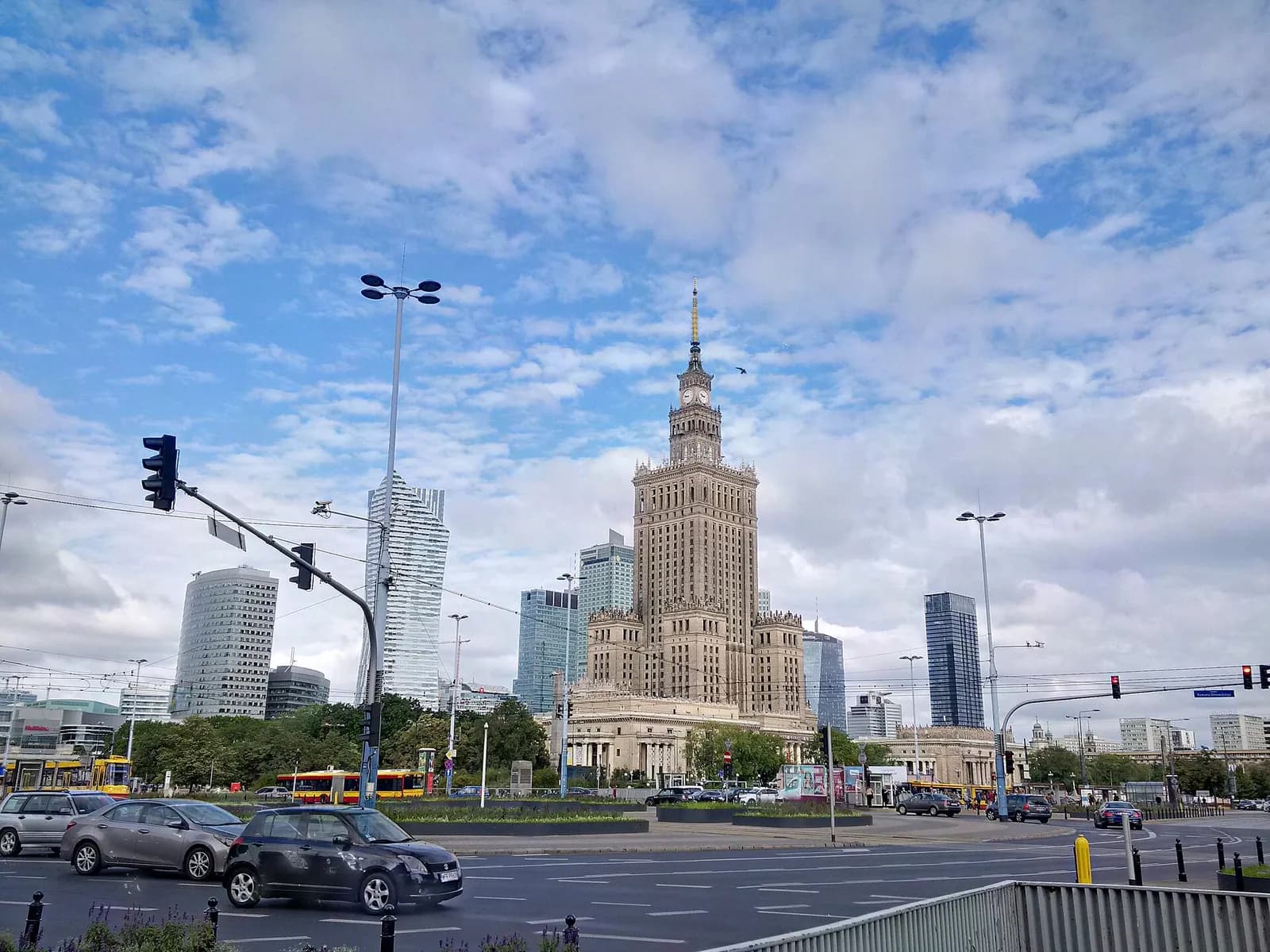 A grand, tall building with a pointed spire and intricate architecture stands prominently against a cloudy sky, surrounded by other modern skyscrapers and a busy street with cars and traffic lights in the foreground.